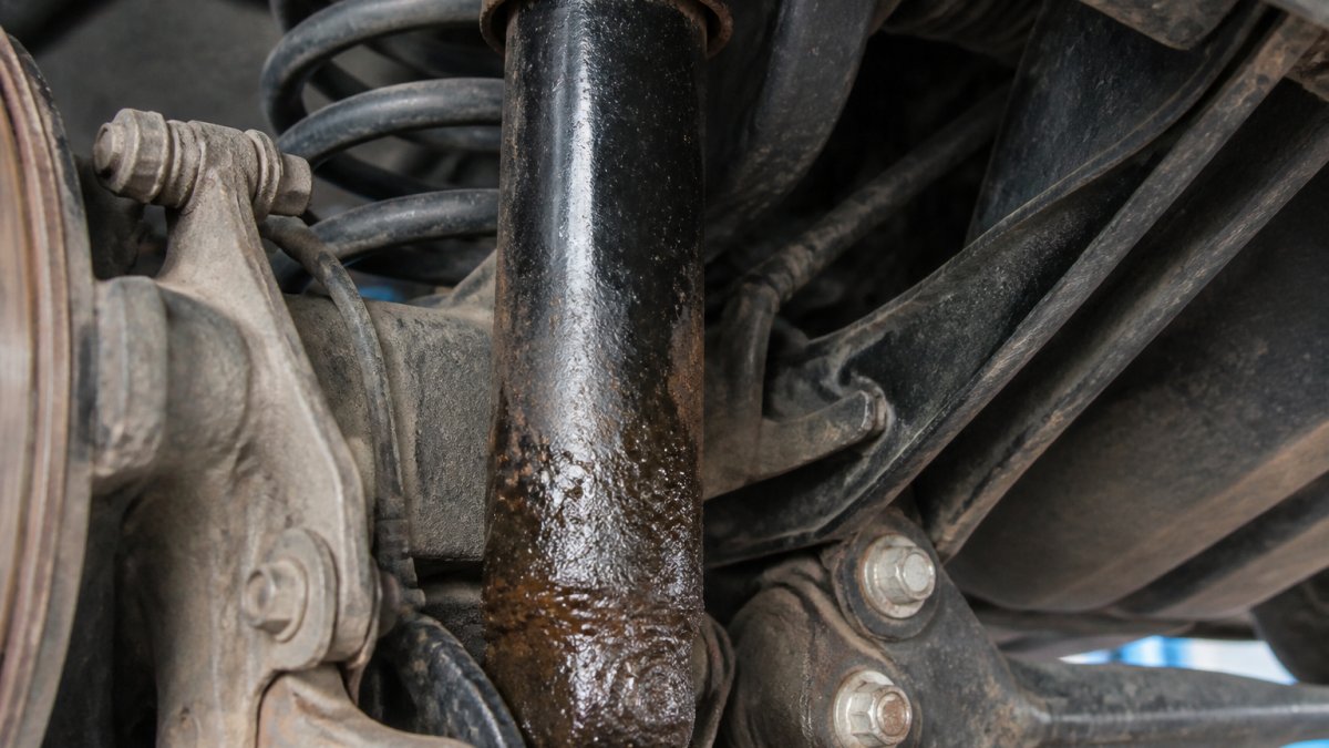 Close-up of vehicle suspension component showing oil seepage and corrosion on shock absorber body in workshop setting with bright task lighting