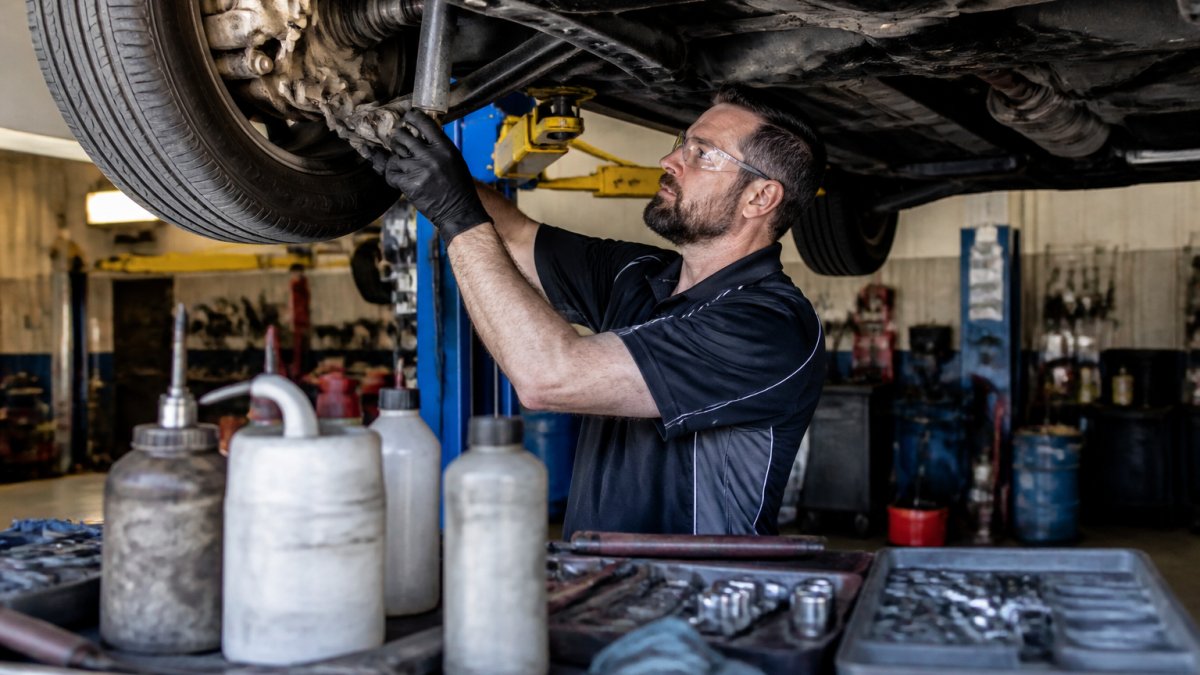 Professional mechanic inspecting brake components and suspension parts under a vehicle lifted on a service bay hoist, with diagnostic tools and fluid containers visible nearby